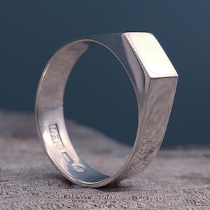 May include: A silver signet ring with a rectangular face. The ring has a polished finish and a smooth band. The ring is shown on a wooden surface with a dark blue background. The ring is stamped with the word "DEN".