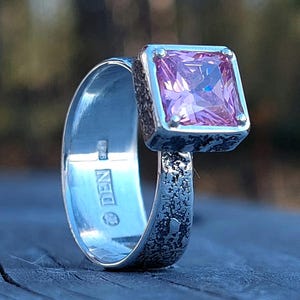 May include: A sterling silver ring with a square, light purple gemstone. The ring has a textured band and setting, with a polished interior. The gemstone is set in a square bezel setting. The ring is a piece of jewelry.