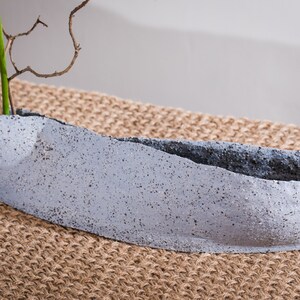 以下が含まれることがあります： ざらざらとした質感の薄い灰色の陶器の花瓶。花瓶は楕円形で、開口部が広い。茶色の織り目の表面の上に置かれている。