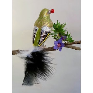 Op de afbeelding: Decoratief glazen vogelornament met een groen lichaam, zilveren en zwarte accenten en een rode bes. De vogel heeft een lange staart van zwarte en witte veren en zit op een tak met paarse bloemen en groene bladeren. Een unieke vakantiedecoratie.