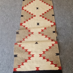 May include: A rectangular runner rug with a geometric pattern. The rug features a cream-colored central field with diamond shapes in red and black. The sides have alternating sections of tan and cream, with black tassels at each end.
