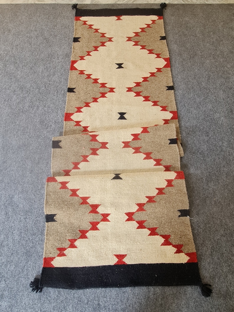 May include: A rectangular runner rug with a geometric pattern. The rug features a cream-colored central field with diamond shapes in red and black. The sides have alternating sections of tan and cream, with black tassels at each end.