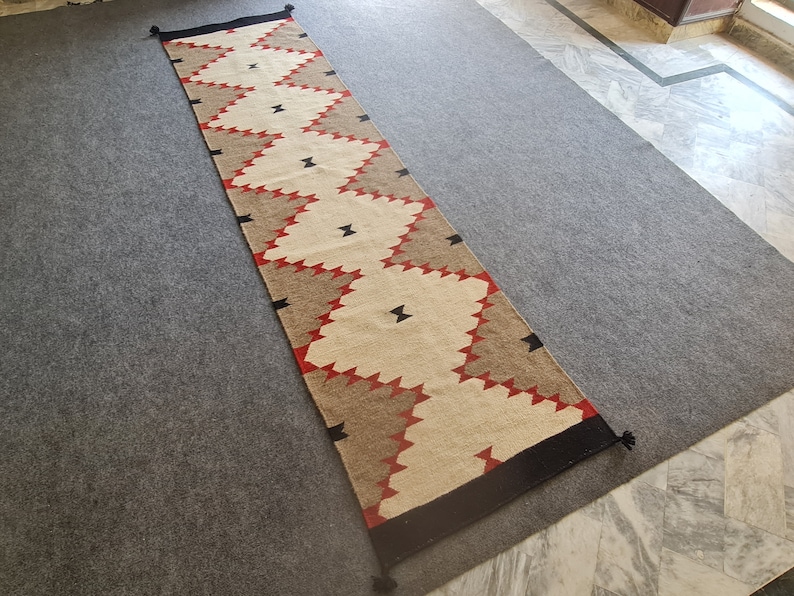 May include: A long, rectangular runner rug with a geometric pattern. The rug features alternating sections of beige, brown, and red zigzags, with black accents and tassels at each end. The rug is placed on a gray surface.