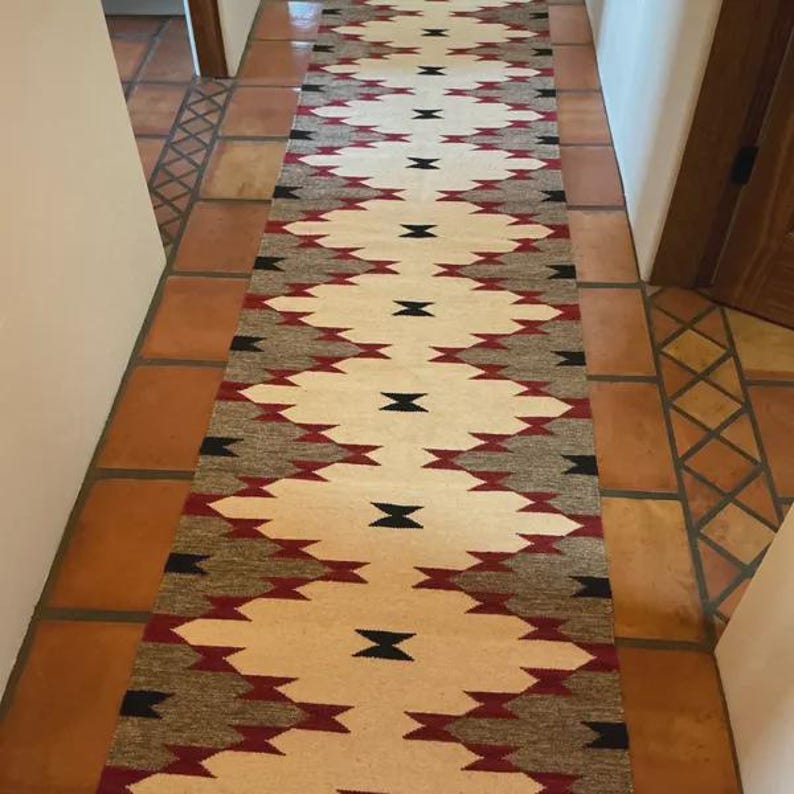 May include: A long, rectangular rug with a geometric pattern in cream, red, black, and gray. The rug is placed in a hallway with terracotta tile flooring. The design features repeating diamond shapes.