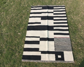 Puede incluir: Alfombra geométrica con patrón en blanco y negro con una pequeña franja roja y un cuadrado blanco y negro en el centro.
