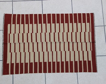 Puede incluir: Una alfombra rectangular con un patrón geométrico de rayas rojas y beige alternas. Las rayas están formadas por rectángulos verticales.