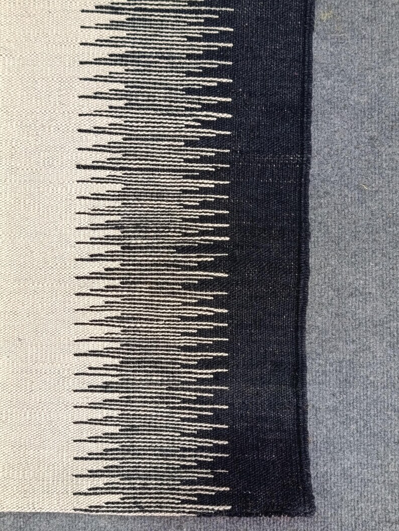 May include: A black and white woven rug with a geometric pattern. The rug has a fringe border and is made of natural fibers.