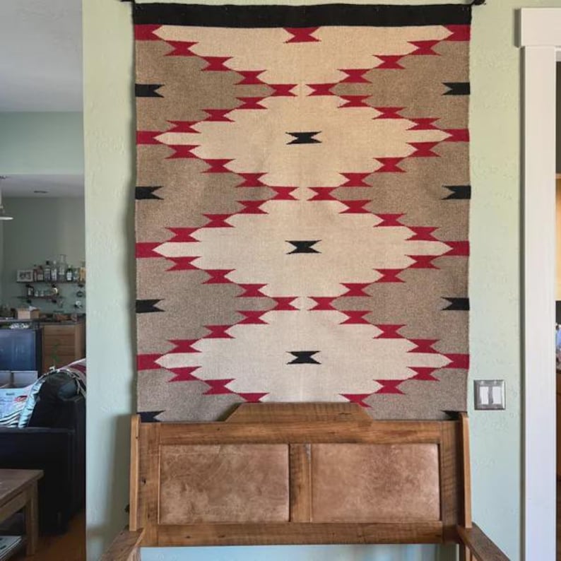May include: A woven wall hanging with a geometric pattern in shades of beige, gray, red, and black. The design features diamond shapes and zigzags. The wall hanging is displayed above a wooden headboard.