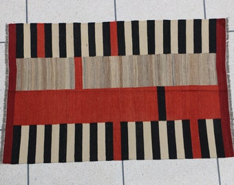 Puede incluir: Alfombra rectangular tejida con un patrón geométrico. La alfombra presenta rayas verticales negras y crema alternas, con una sección central de color beige y rojo. Los bordes tienen flecos.