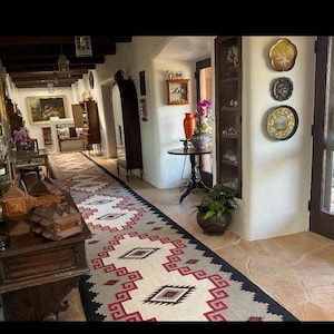 Puede incluir: Un largo pasillo con una alfombra estampada con diseño geométrico en rojo, negro y crema. Las paredes son blancas y el suelo es de baldosas. Se ven muebles antiguos y objetos decorativos, incluyendo cuadros y platos decorativos.