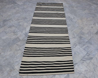 May include: A long, narrow, white and black striped rug. The rug has a simple design with alternating black and white stripes.