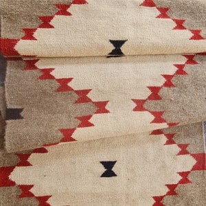 May include: A folded, woven runner rug with a geometric pattern. The rug features a cream-colored central field with diamond shapes outlined in red and bordered by a gray gradient. Black accent shapes are also present.