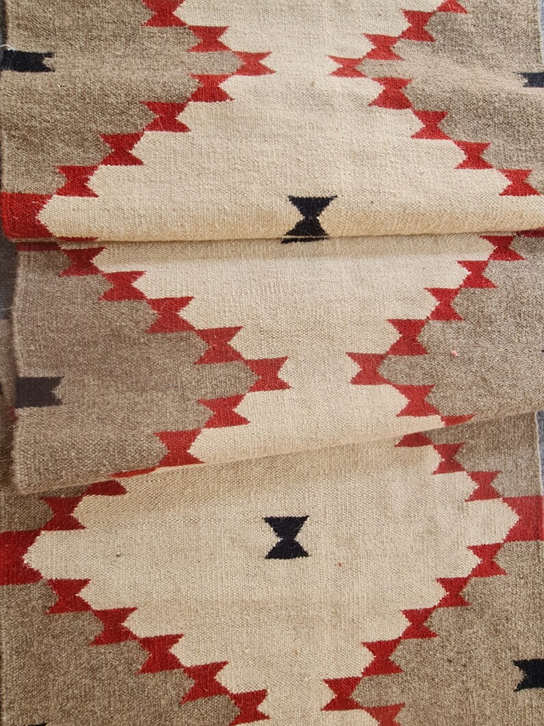 May include: A folded, woven runner rug with a geometric pattern. The rug features a cream-colored central field with diamond shapes outlined in red and bordered by a gray gradient. Black accent shapes are also present.