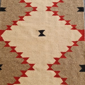 May include: Woven rug with a geometric pattern. The rug features a central beige field with a diamond pattern in red, brown, and black. The rug is rectangular and appears to be made of natural fibers.