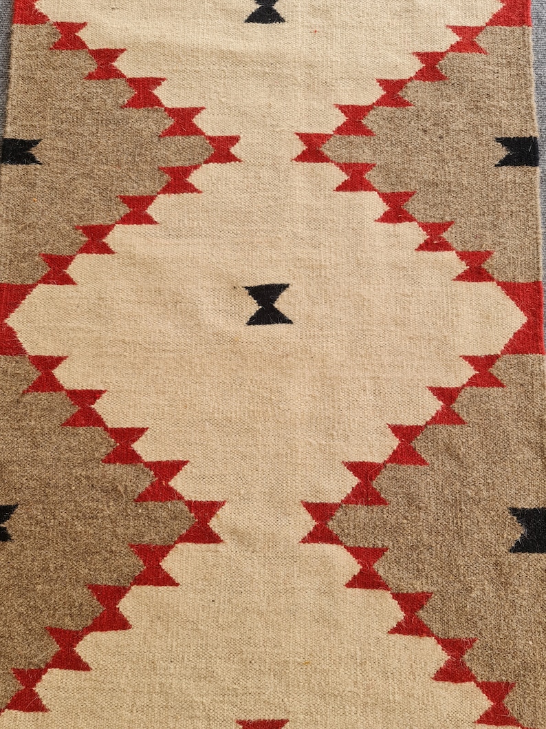 May include: Woven rug with a geometric pattern. The rug features a central beige field with a diamond pattern in red, brown, and black. The rug is rectangular and appears to be made of natural fibers.