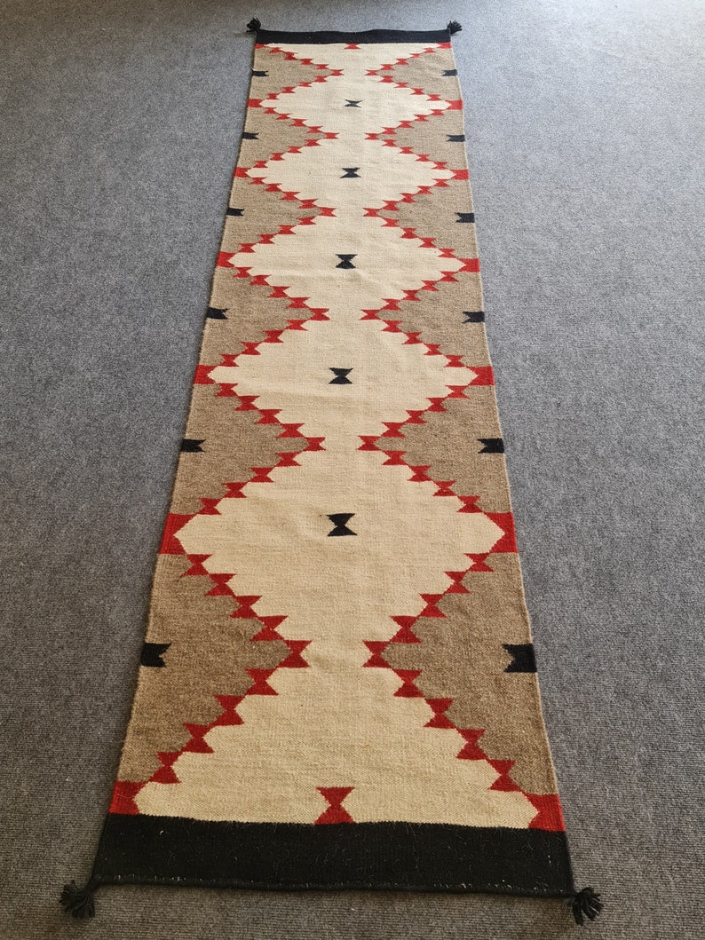 May include: A long, rectangular runner rug with a geometric pattern. The rug features a central diamond design in cream, brown, and red, with black accents. The ends are black with tassels. The rug is made of woven material.