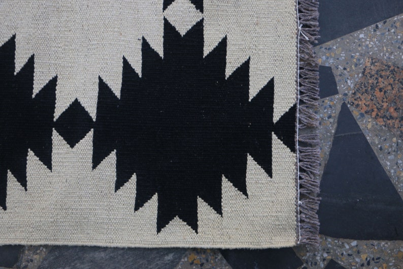 Puede incluir: Alfombra de patr&oacute;n geom&eacute;trico en blanco y negro con flecos en el lado derecho.