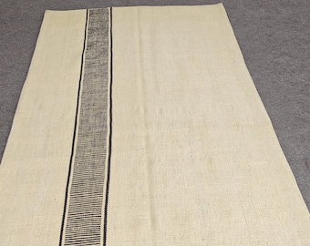 May include: A rectangular, cream-colored rug with a central vertical stripe in dark brown. The stripe is bordered by thin black lines. The rug is made of a woven material, and the background is a neutral gray.