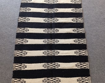 May include: A rectangular black and cream-colored rug with horizontal stripes and decorative geometric patterns. The rug has a fringed edge and is suitable for use as a floor covering or decorative accent.
