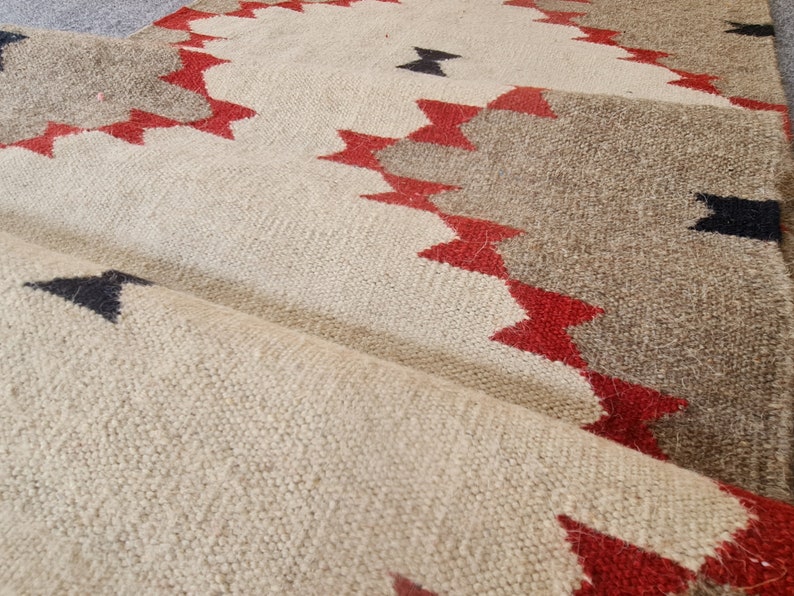 May include: Close-up of a woven rug with a geometric pattern. The rug features a cream-colored background with brown and red zigzag designs. Black arrow shapes are scattered across the surface. The rug is folded over, revealing its texture.