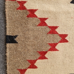 May include: Close-up of a woven textile with a geometric pattern. The design features alternating bands of beige, brown, and red, with black accents. The texture of the fabric is visible, showcasing the craftsmanship.