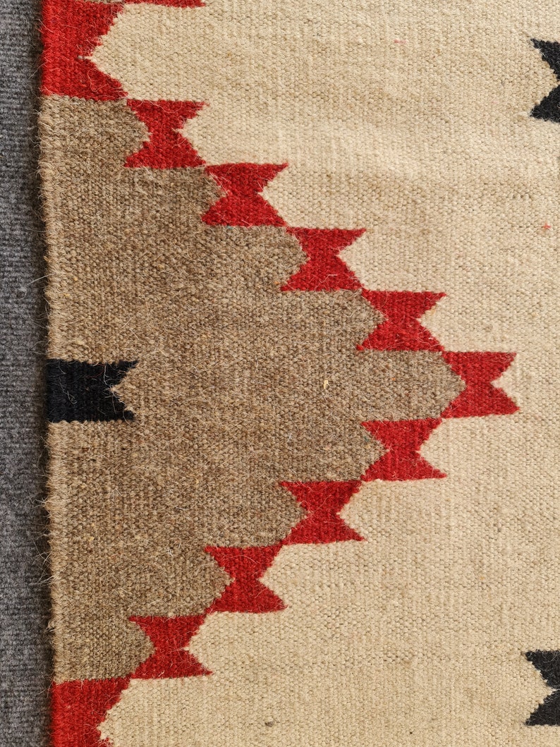 May include: Close-up of a woven textile with a geometric pattern. The design features alternating bands of beige, brown, and red, with black accents. The texture of the fabric is visible, showcasing the craftsmanship.