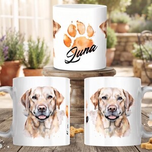 Könnte beinhalten: Ein weißer Keramikbecher mit einer Aquarellillustration eines gelben Labrador Retrievers auf der Vorderseite und einem Pfotenabdruck mit dem Namen Juna in schwarzer Schreibschrift auf der Rückseite, ergänzt durch braune und beige Akzente.