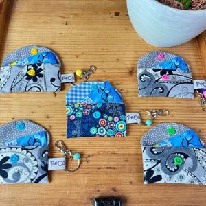 Könnte beinhalten: Eine Sammlung von Stoff-Münzbörsen mit Schlüsselanhängern. Die Börsen weisen verschiedene Muster auf, darunter Blumen-, Paisley- und geometrische Designs in Blau, Grau und Weiß. Jede Börse hat einen Knopfverschluss und ein kleines Etikett mit der Aufschrift "PeCa".
