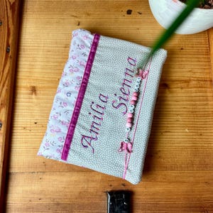 Könnte beinhalten: Ein personalisiertes Babybuch mit dem Namen "Amilia Sienna" in lila Schrift gestickt. Das Buch hat ein Blumenmuster und einen rosa Bandakzent. Ein Perlenanhänger mit dem Datum "07 2020" hängt am Buch.