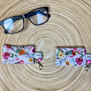 Könnte beinhalten: Zwei kleine rechteckige Stoffbeutel mit Blumenmuster in Rosa-, Gelb- und Grüntönen, jeweils mit grünem Druckknopf, weißem Etikett mit der Aufschrift PeCa, einer rosa Perle und einem Metallkarabiner. Darüber befinden sich eine schwarze Brille.