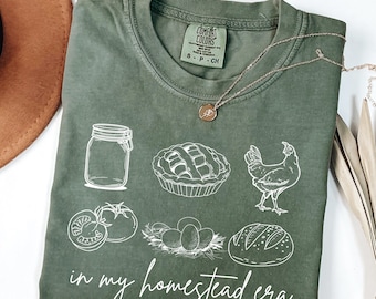 May include: Olive green t-shirt with white illustrations of a jar, pie, chicken, tomatoes, eggs, and bread. The text "in my homestead era" is printed in white script. A gold necklace is draped across the shirt.