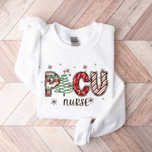 May include: White PCU nurse sweatshirt. The letters "PCU" are in a festive design with red and green patterns, and a Christmas tree graphic. The word "nurse" is below the letters, with snowflakes and stars.