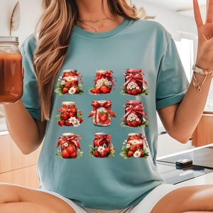 May include: A teal blue t-shirt with a graphic of nine jars filled with red strawberries. Each jar has a red ribbon and white flowers around the base.