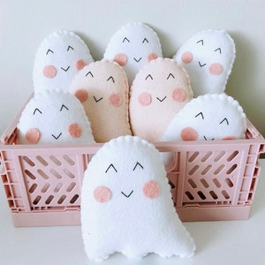 May include: A pink crate filled with eight white and beige ghost-shaped plush toys. The ghosts have embroidered eyes and mouths and pink felt cheeks.