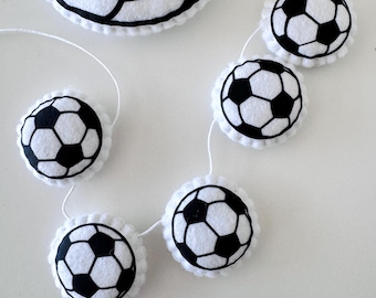 May include: A white felt garland featuring black and white soccer ball designs. The garland includes multiple soccer ball shapes connected by a white string. The soccer balls have black pentagon and hexagon patterns.