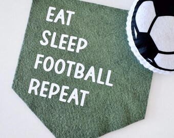 May include: A green felt banner with the words "EAT SLEEP FOOTBALL REPEAT" in white block letters. A white and black soccer ball is to the right of the banner. The banner is hanging from a white string.