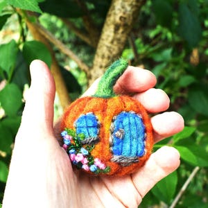 May include: A needle-felted orange pumpkin ornament featuring a green stem, a blue arched door, a blue window, and small pink and blue floral accents made of beads. The pumpkin has a textured, fuzzy surface and is round in shape.