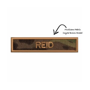 Puede incluir: Un parche rectangular con el nombre "REID" en letras en relieve. El parche presenta un fondo de tela multicam con un borde de hilo marrón coyote. También está presente el texto "Multicam Fabric Coyote Brown thread".