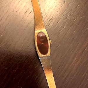 Può includere: Un orologio Seiko Quartz color oro con un cinturino in metallo sottile e testurizzato e una cassa ovale. Il quadrante è marrone scuro con un piccolo accento a ore dodici, lancette dorate e un bordo perlato attorno al quadrante.