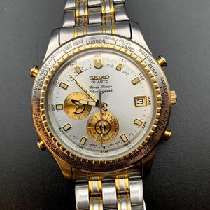May include: A Seiko Quartz World Timer Chronograph watch with a silver and gold-tone metal link band. The white dial features gold-tone hour markers, three sub-dials, a date window, and a rotating bezel printed with global city names.