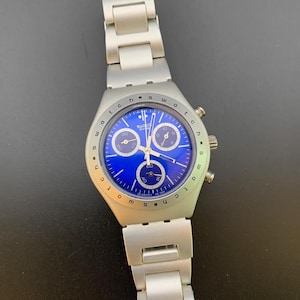May include: A silver-toned metal chronograph wristwatch featuring a vibrant blue dial with three sub-dials. The bezel is marked with the word Swatch repeated around the perimeter. The watch includes a matching silver-toned link bracelet band.