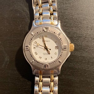 May include: A silver and gold-toned wristwatch with a white face. The watch features Roman numerals, a date display, and a textured bezel. The watch band is a combination of silver and gold links.