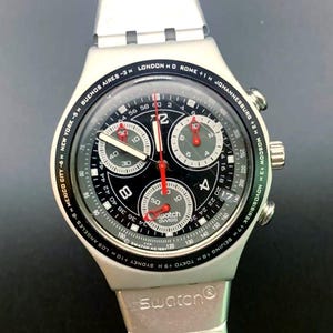 May include: A silver-toned Swatch chronograph wristwatch featuring a black dial with three sub-dials, white Arabic numerals, and red hands. The bezel displays global city names and time zone offsets, and the metal link bracelet has black accents.