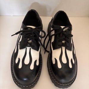 May include: A pair of black and white lace-up shoes featuring a prominent flame pattern design. The shoes have a rounded toe, black laces, and a thick, lugged sole with visible white stitching along the edge of the base, creating a bold contrast.