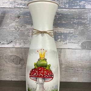 May include: White ceramic vase featuring a frog wearing a gold crown perched on a red and white mushroom. The vase is adorned with a brown twine bow and a silver charm inscribed with "Good Vibes Only".