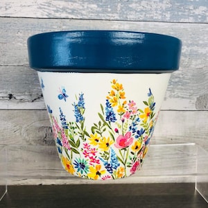 May include: A white terracotta flower pot with a blue rim and a floral design featuring pink, yellow, blue, and green flowers.