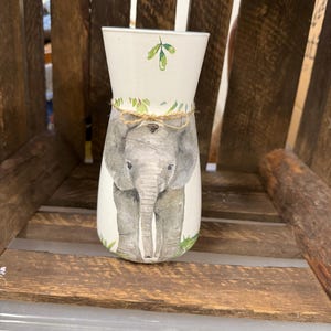 Puede incluir: Jarrón de cerámica blanco con una ilustración de acuarela de un elefante y hojas verdes. El jarrón tiene un lazo de cuerda marrón alrededor de la parte superior.