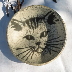 Puede incluir: Un cuenco redondo de cerámica con un diseño de cara de gato pintado a mano en negro sobre un fondo color crema. La superficie tiene un acabado de esmalte craquelado distintivo, creando una red de líneas finas por todo el interior.