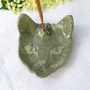 May include: A sage green ceramic incense holder shaped like a cat's face. The holder has a crackle glaze finish and a hole for the incense stick. A lit incense stick is inserted into the holder, with a pink string visible. The background is a white cloth.