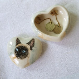 Op de afbeelding: Hartvormige keramische sieradenschaal met een Siamees kattenontwerp en een bijpassend deksel. De schaal bevat gouden oorbellen. De kat heeft blauwe ogen en donkerbruine aftekeningen. De schaal is wit met een lichtbruine en blauwe rand.